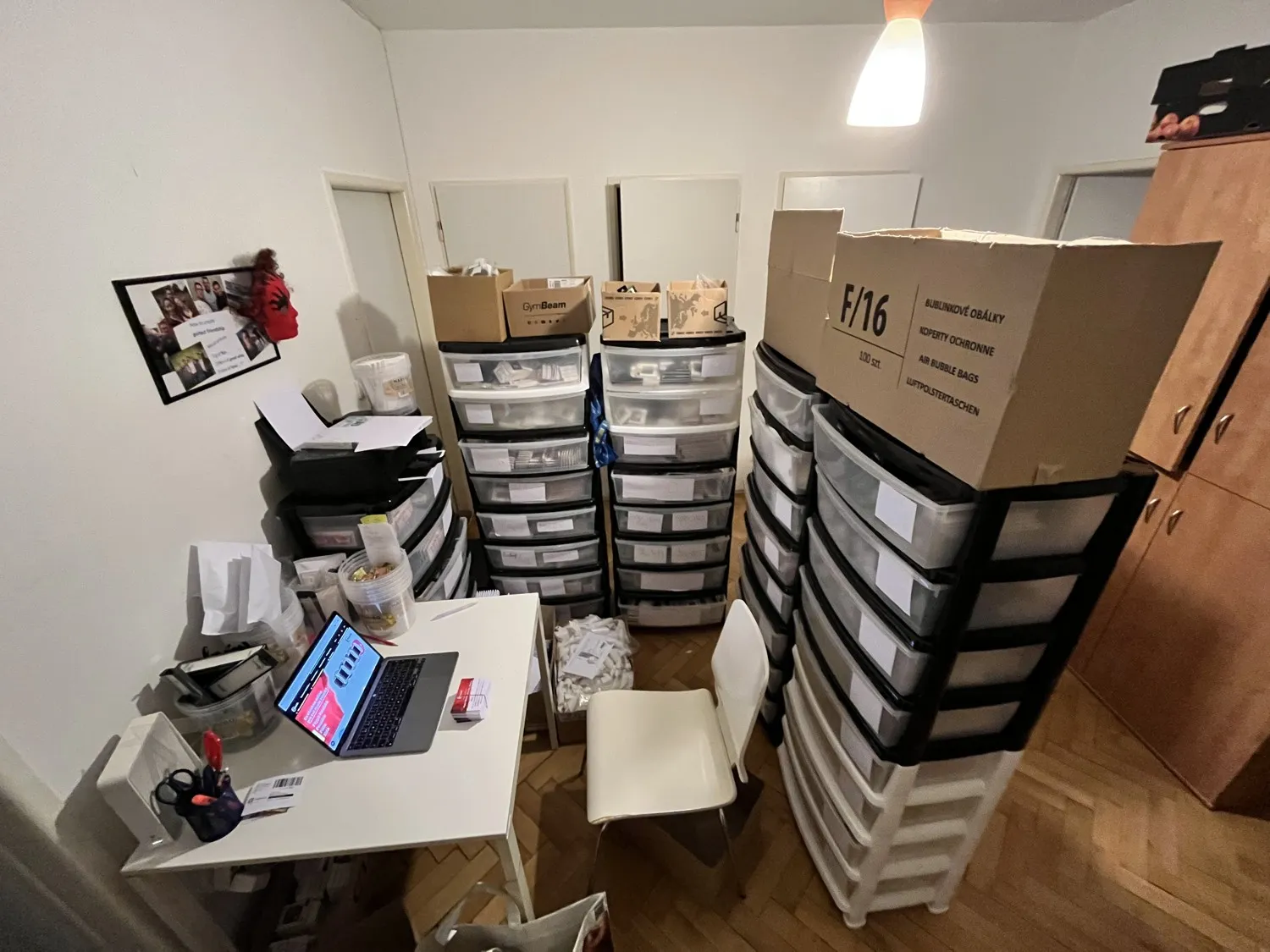 Pokoj v bytě přeměněný ve sklad e‑shopu: stohy plastových šuplíků, krabice s obálkami a balicí stůl